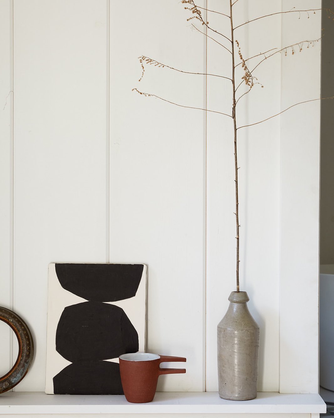 Still life of abstract art, mug, dried branch in bottle, and frame against a white paneled wall create a minimalist interior.