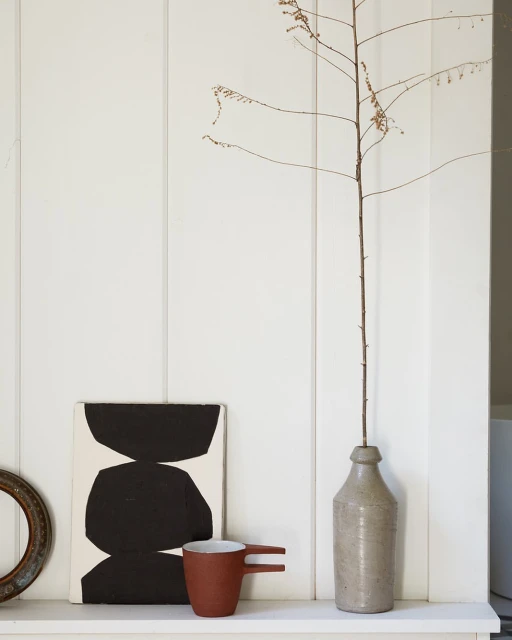 Still life of abstract art, mug, dried branch in bottle, and frame against a white paneled wall create a minimalist interior.