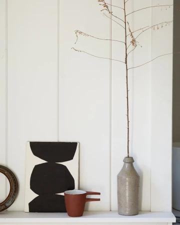 Still life of abstract art, mug, dried branch in bottle, and frame against a white paneled wall create a minimalist interior.