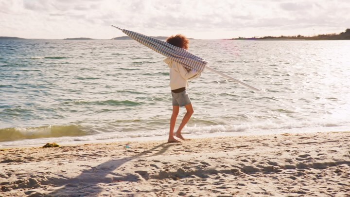 Young woman carries a blue and white striped beach umbrella on a sandy beach with the ocean and sky in the background on a sunny day.