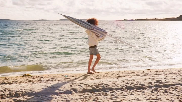Young woman carries a blue and white striped beach umbrella on a sandy beach with the ocean and sky in the background on a sunny day.