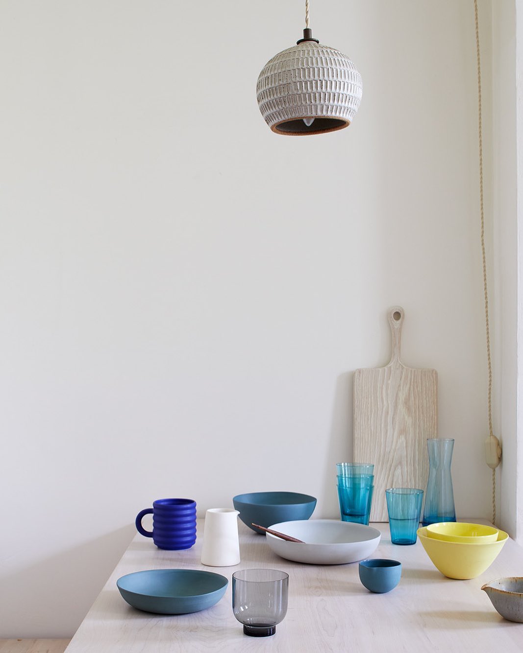 Still life of kitchenware on a light wood table, including ceramic bowls, mugs, glasses, a cutting board, and a pendant light fixture.