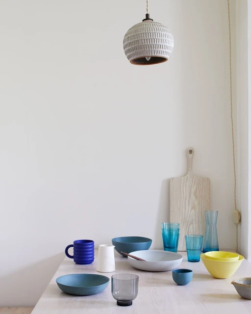 Still life of kitchenware on a light wood table, including ceramic bowls, mugs, glasses, a cutting board, and a pendant light fixture.