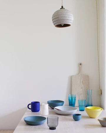 Still life of kitchenware on a light wood table, including ceramic bowls, mugs, glasses, a cutting board, and a pendant light fixture.
