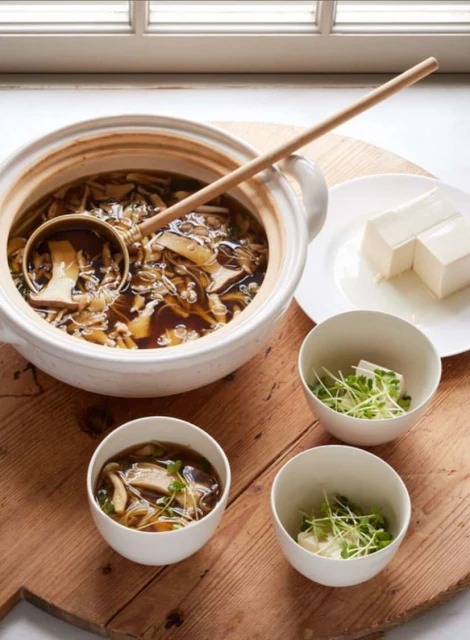 Overhead shot of mushroom soup in a ceramic pot with a ladle, served with tofu and sprouts on a wooden board; food photography.
