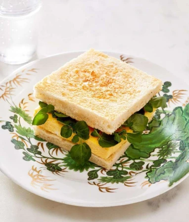 Close-up of a square egg sandwich on a decorative plate, topped with fresh green watercress; white bread and clean presentation.