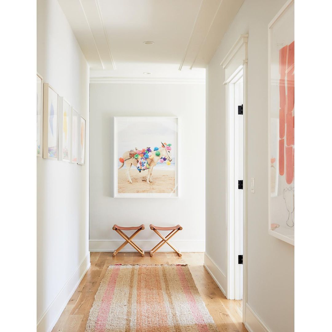 Hallway interior with a framed art print of an animal decorated with colorful pompoms hanging above two wooden folding stools.