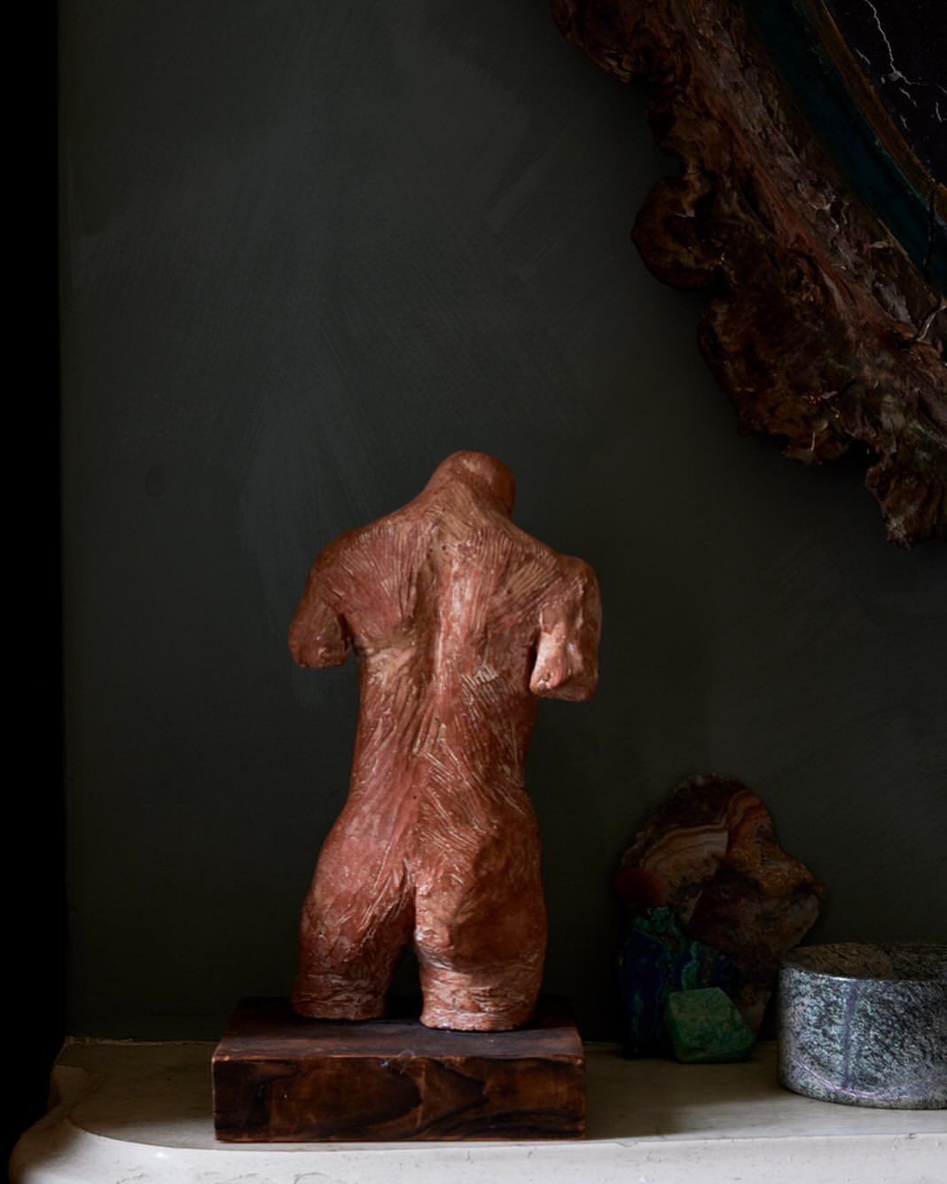 Back view of a nude torso sculpture in copper tones, displayed indoors with rocks and decorative wall hanging in a dark ambient setting.