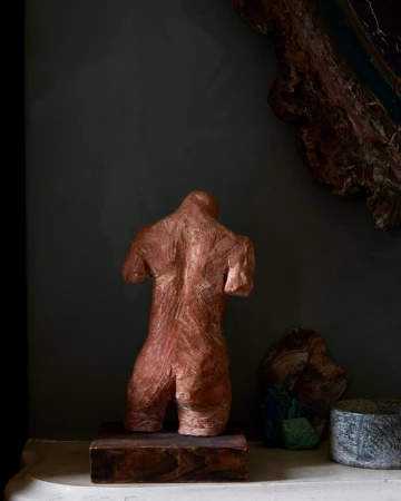Back view of a nude torso sculpture in copper tones, displayed indoors with rocks and decorative wall hanging in a dark ambient setting.