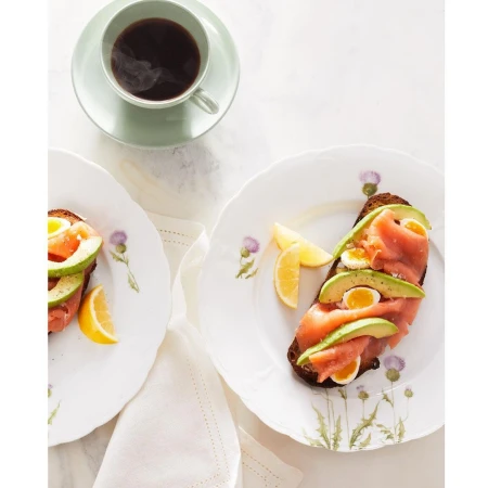 Overhead shot of two plates of toast topped with salmon, avocado, and egg slices, accompanied by lemon wedges and a cup of dark coffee.