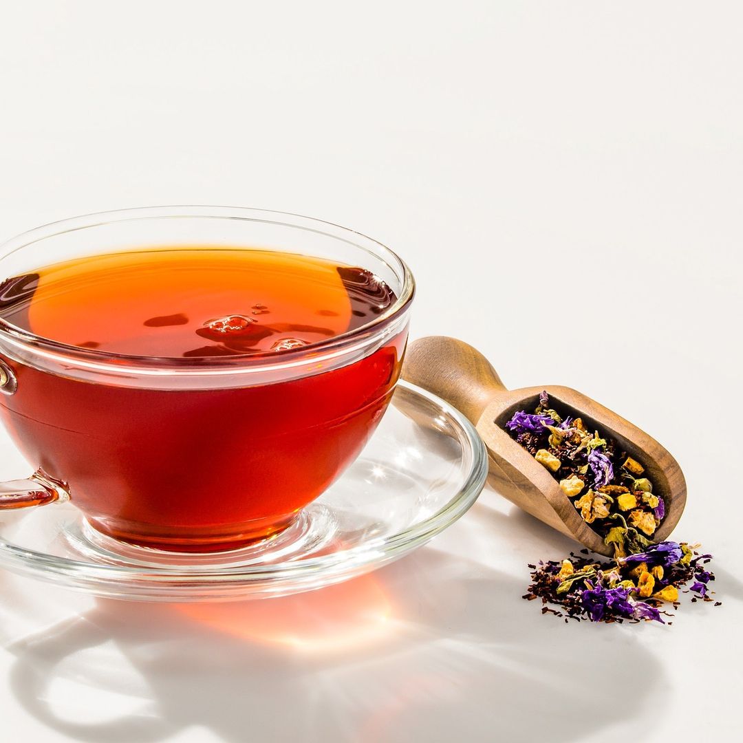 A clear glass teacup filled with amber liquid sits on a saucer next to a wooden scoop with colorful tea leaves, all on a white surface.