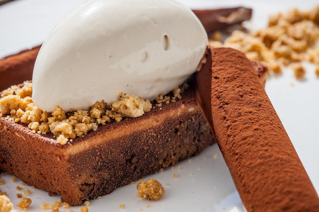 A dessert featuring a rectangular slice of cake topped with white ice cream and sprinkled with crumbs, accompanied by chocolate-covered sticks.