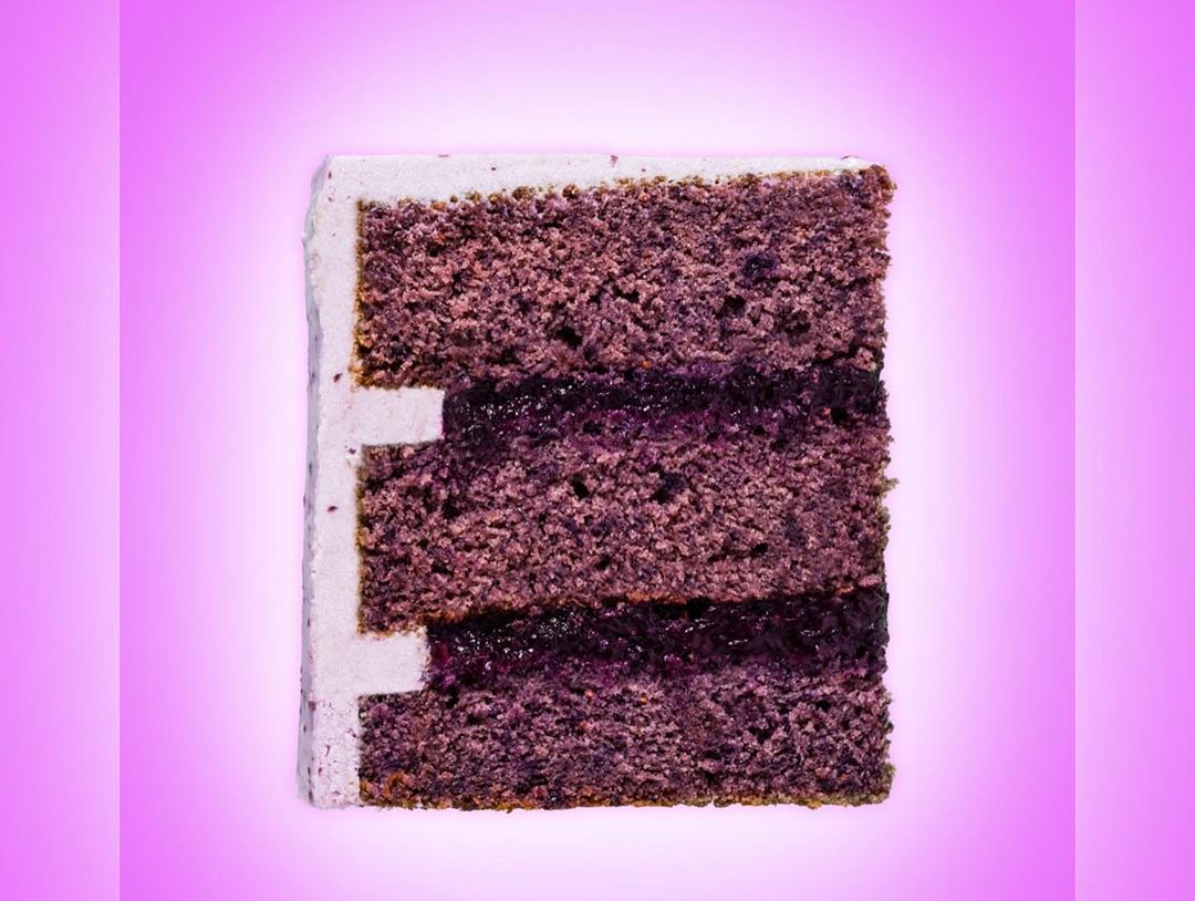 Close-up of a slice of layered cake with frosting, and a dark filling, set against a gradient pink background, food photography.