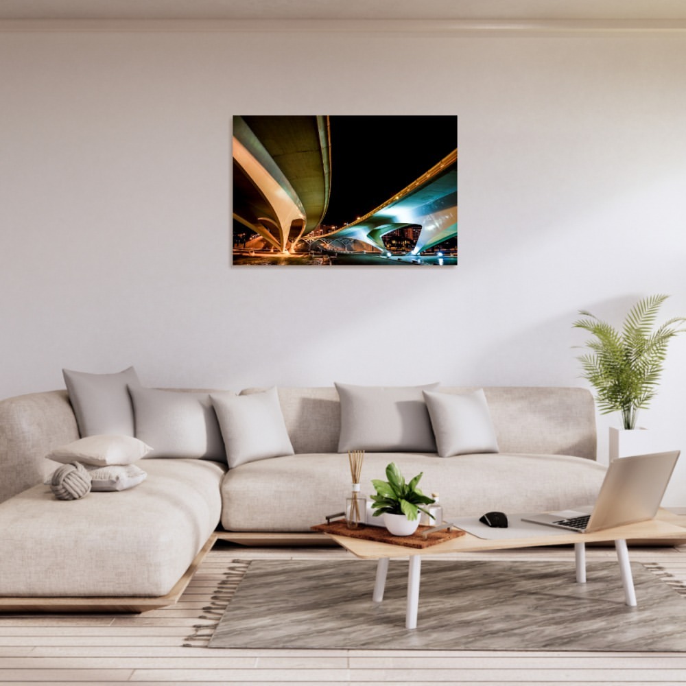 Living room display: couch, pillows, coffee table with laptop, rug, houseplant. Framed art print of a modern bridge at night hangs on wall.