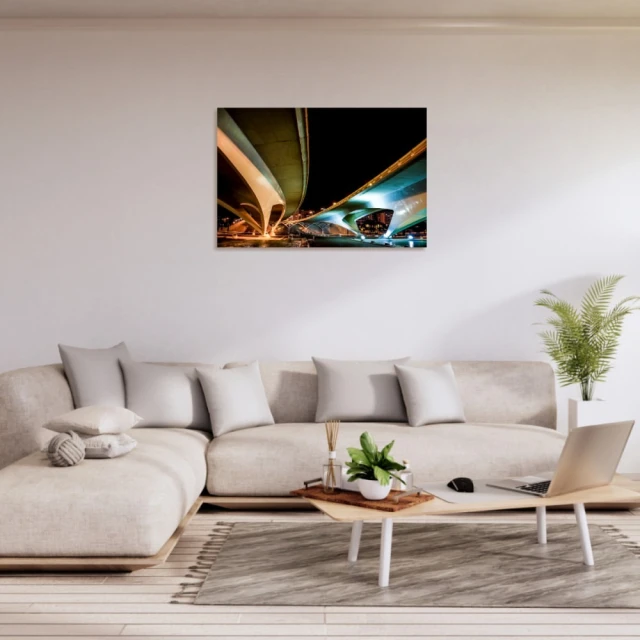 Living room display: couch, pillows, coffee table with laptop, rug, houseplant. Framed art print of a modern bridge at night hangs on wall.