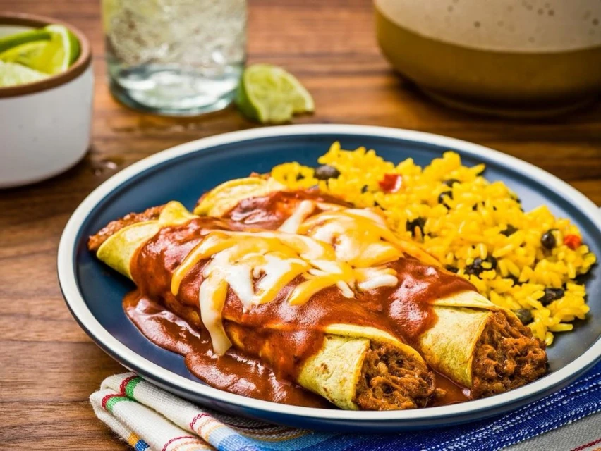 Overhead shot of enchiladas topped with cheese and sauce, served with Mexican rice on a blue plate, set on a wooden table.