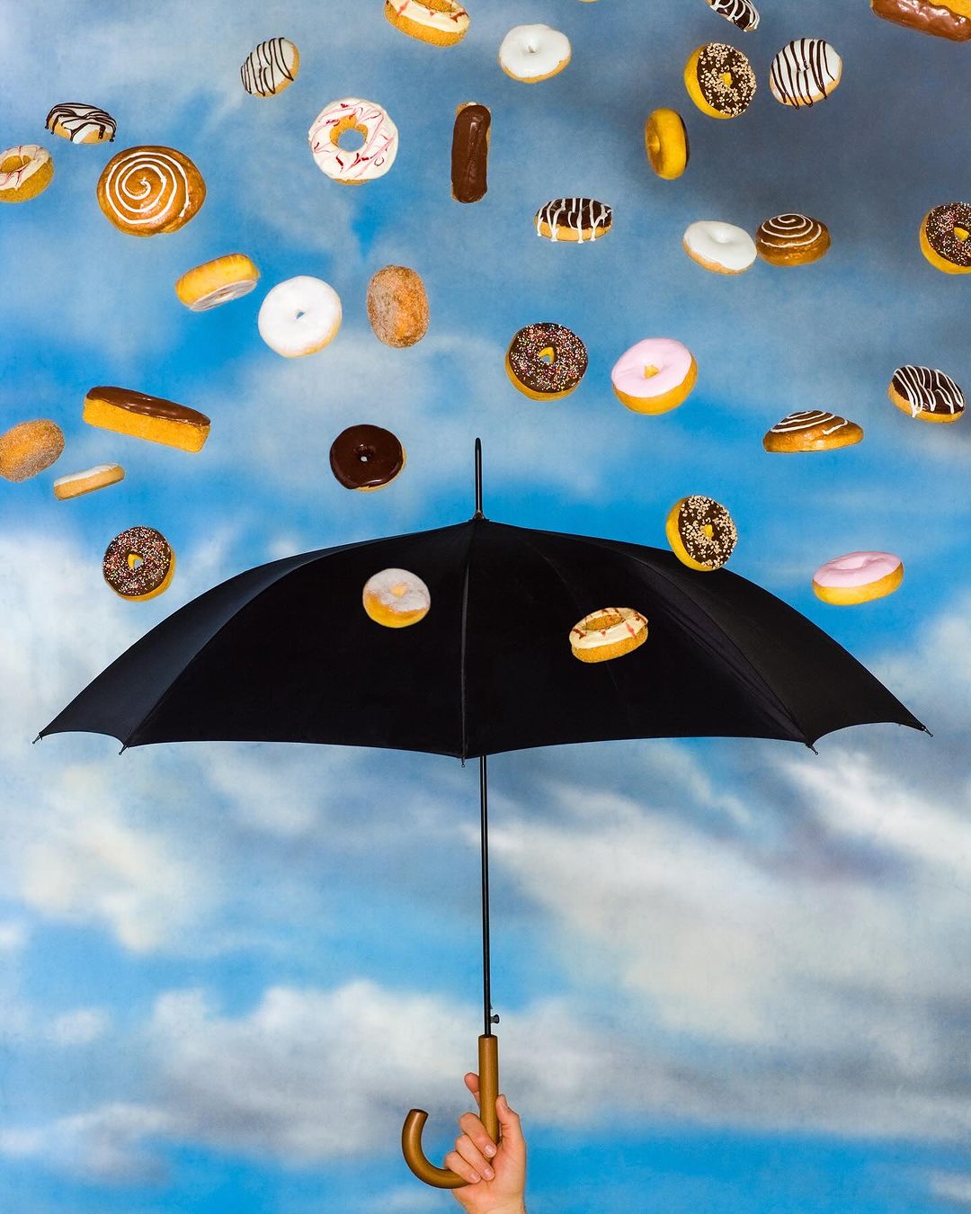 Conceptual image of various types of doughnuts falling from the sky and being caught by a black umbrella against a blue cloudy background.
