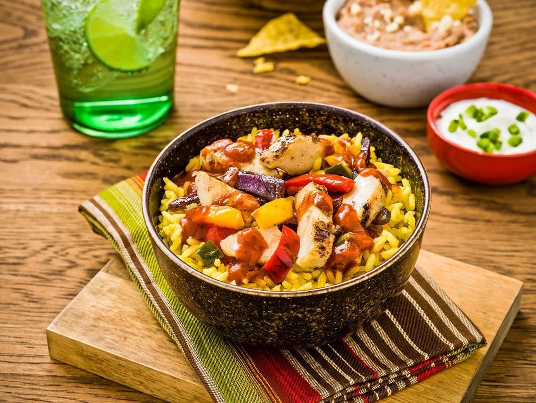 A bowl of rice and chicken with peppers, served on a wooden surface with dips and a drink in the background.