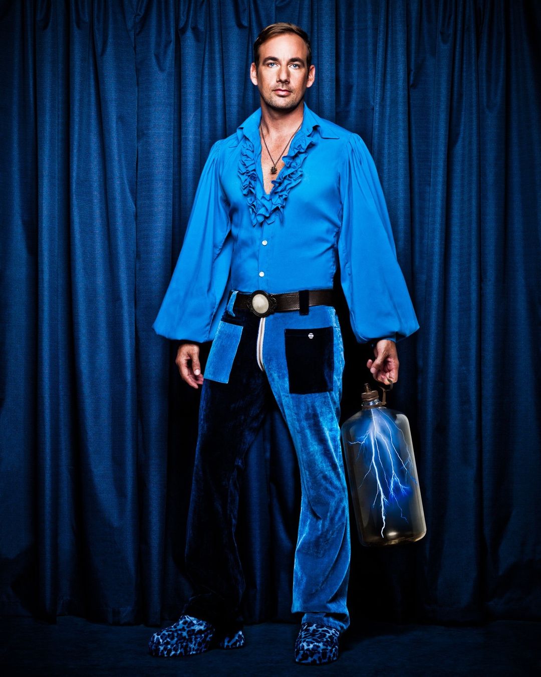 Adult male in blue ruffled shirt and blue velvet pants poses with lightning bottle against blue curtain background in a studio shot.