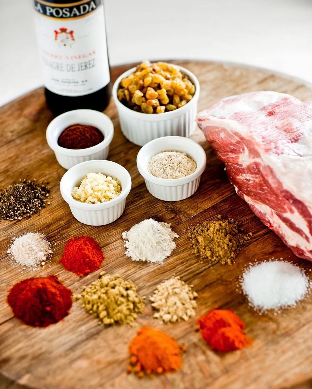 Overhead shot of a pork roast surrounded by small bowls of spices and a bottle of vinegar on a wooden cutting board, food photography.