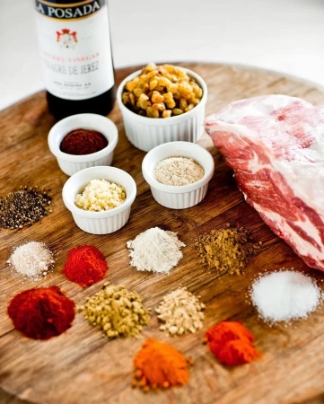 Overhead shot of a pork roast surrounded by small bowls of spices and a bottle of vinegar on a wooden cutting board, food photography.