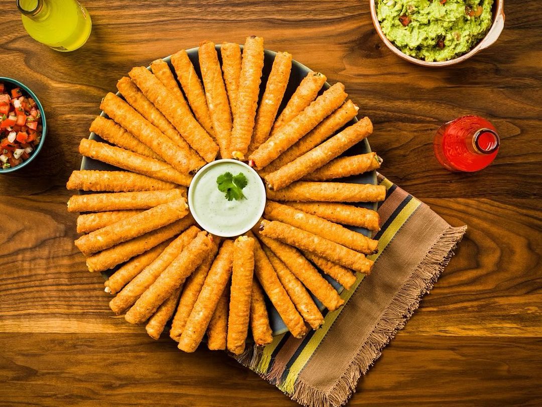 Overhead shot of a platter of fried taquitos with guacamole and salsa dips, and yellow and red bottled sauces on a wood table.