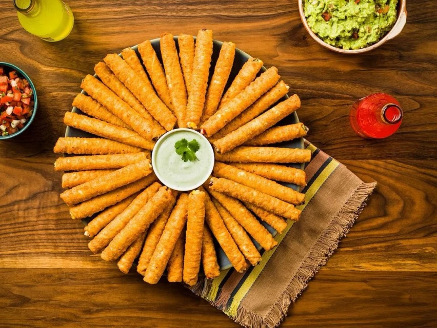 Overhead shot of a platter of fried taquitos with guacamole and salsa dips, and yellow and red bottled sauces on a wood table.