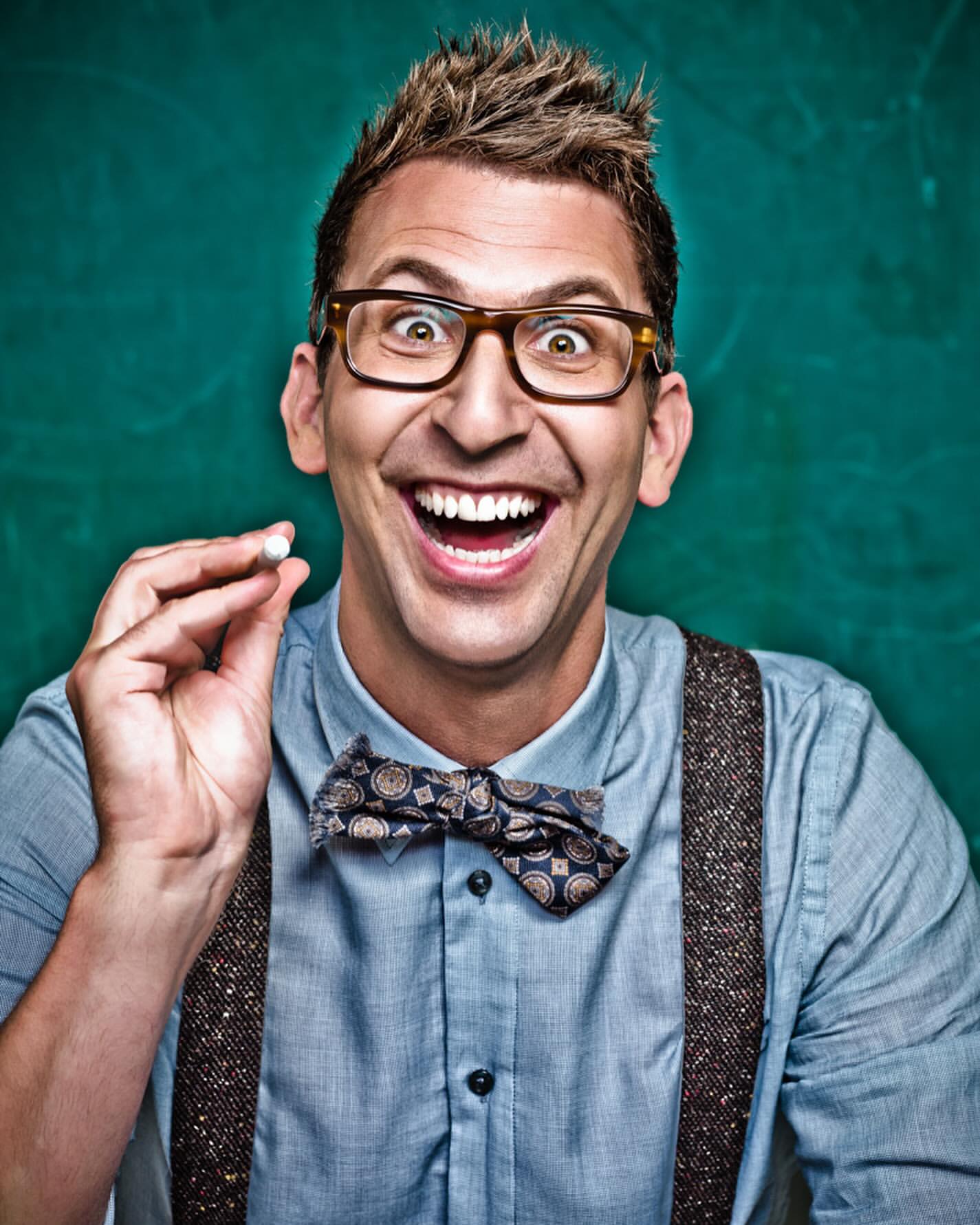 Close-up of a smiling adult male with glasses and a bow tie holding a small white object in his fingers against a green background.