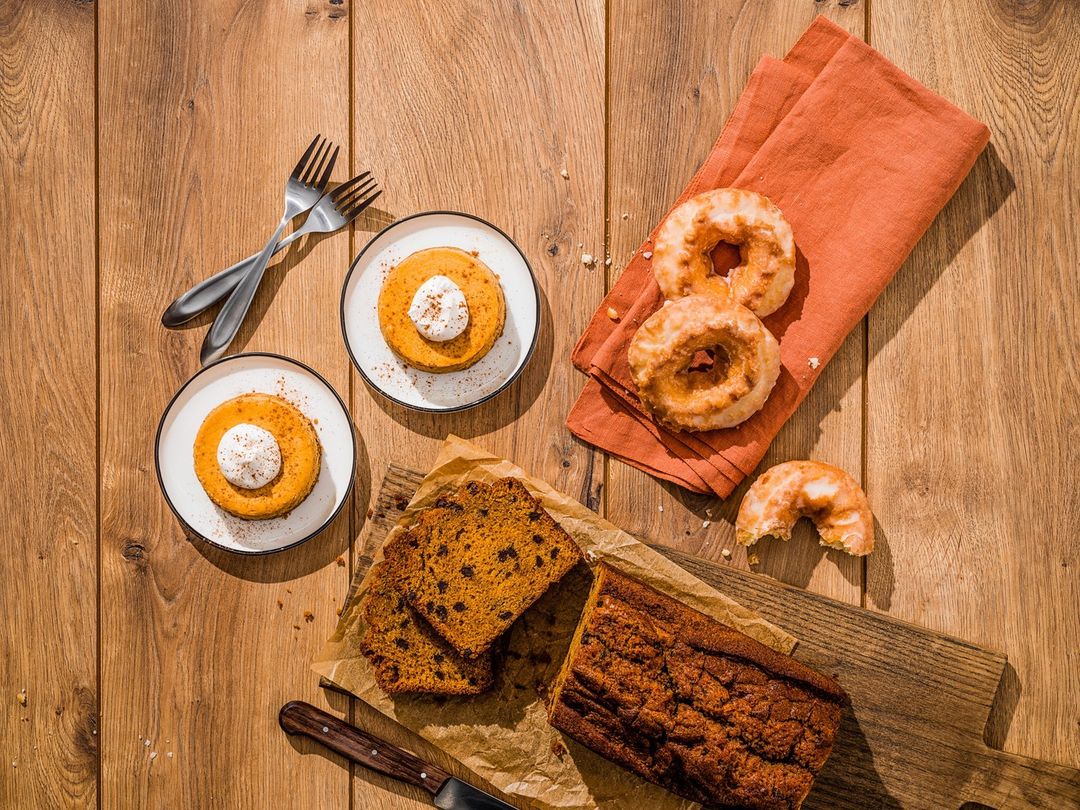 Overhead shot of baked goods, including pumpkin pancakes, cake, and glazed donuts, arranged on a wood table with forks and a linen napkin.