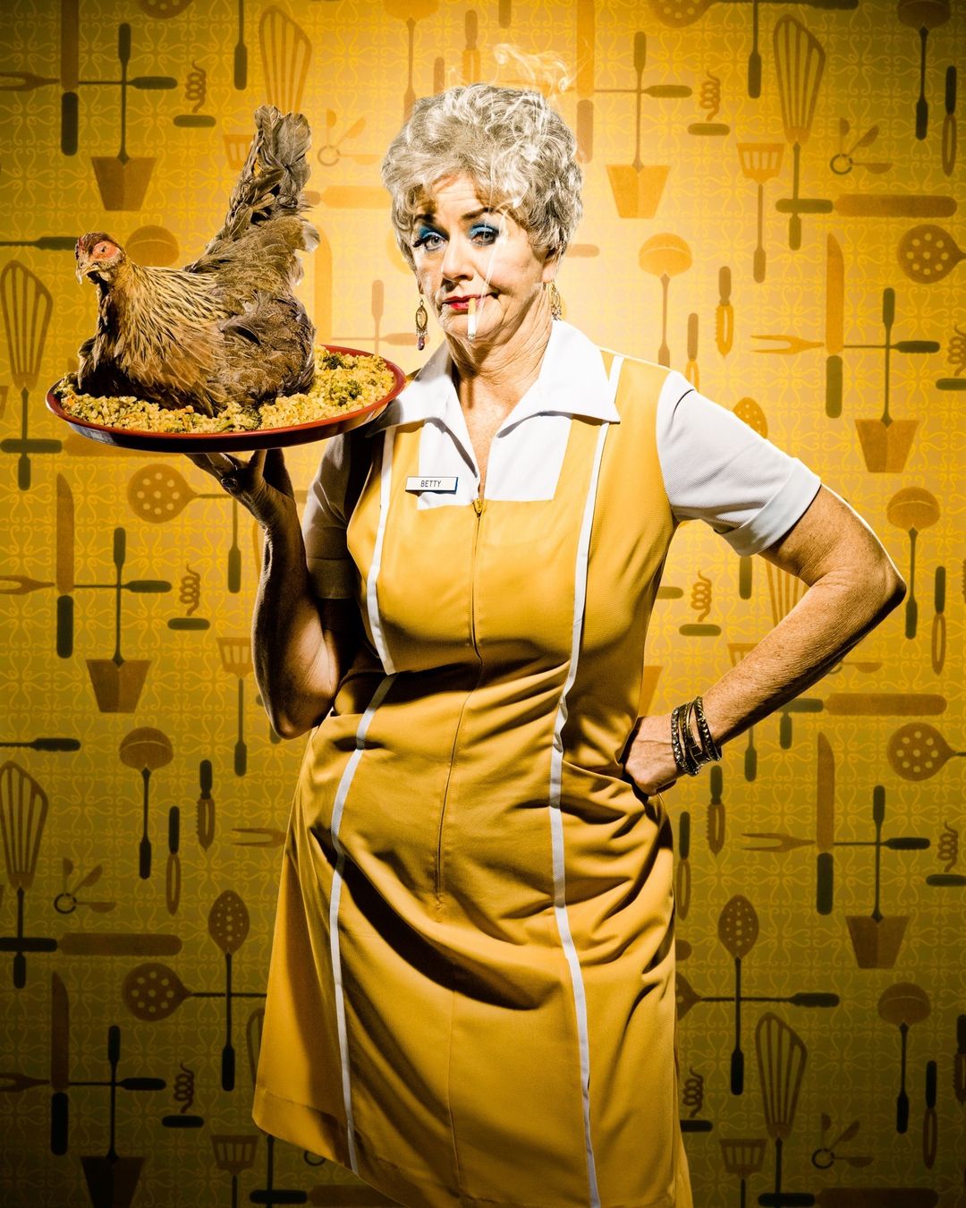 Humorous studio shot of a mature woman in a yellow uniform holding a platter with a raw chicken and kitchen utensils background.
