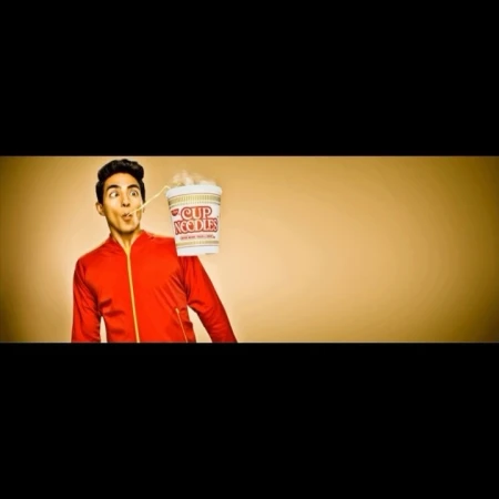 Person in a red jacket eats noodles from a Cup Noodles container on a tan background. The person has a noodle hanging out of their mouth.