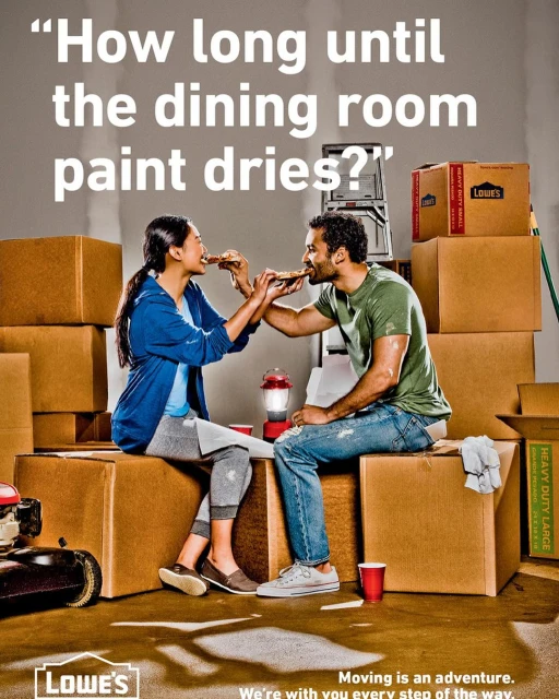 Couple sitting on moving boxes sharing pizza; Lowe's moving ad shows the lighter side of moving day; indoor home shot with moving supplies.