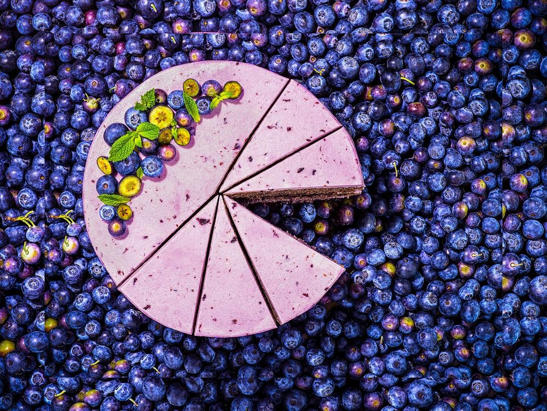 Overhead shot of a lavender-iced cake with blueberries and mint, set against a dense background of blueberries, creating a vibrant food still life.