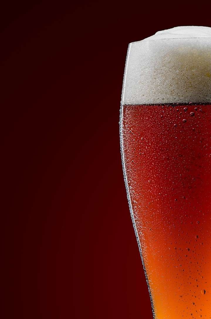 Close-up of a glass of amber beer with a thick white head, condensation on the glass, and a dark red background.