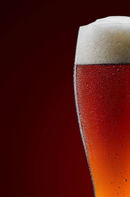 Close-up of a glass of amber beer with a thick white head, condensation on the glass, and a dark red background.