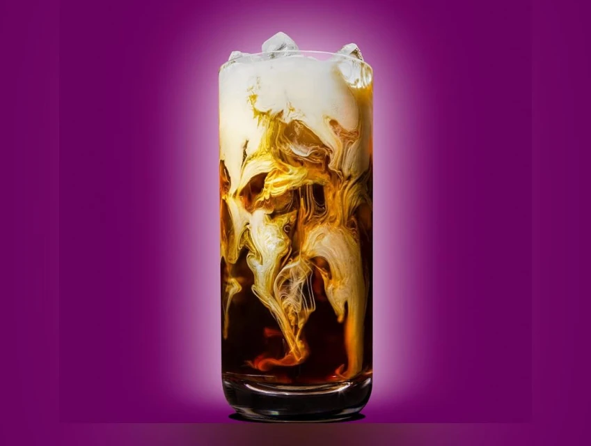 Iced coffee drink in tall clear glass with cream swirled in coffee and ice cubes, set against gradient magenta to white background.