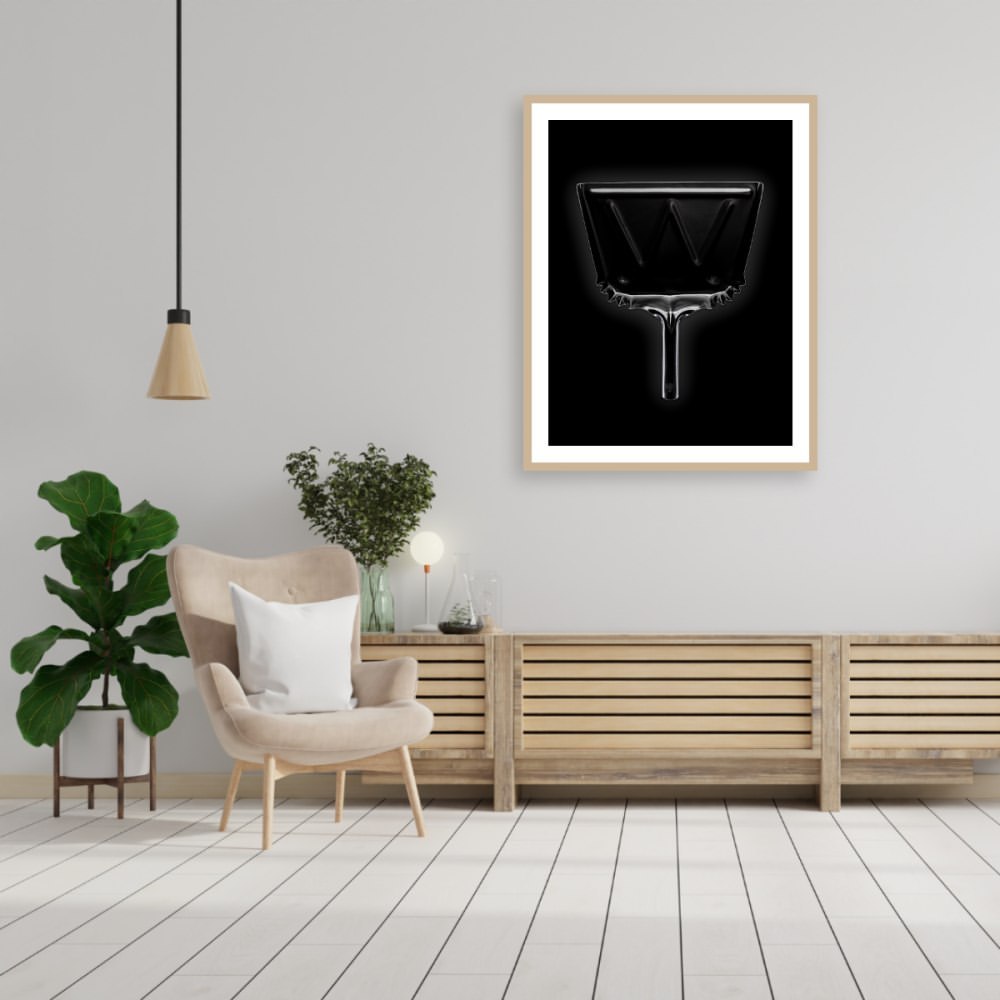 Framed black and white abstract art of inverted trapezoid with interior letter W hangs on wall of staged room with chair and plant.