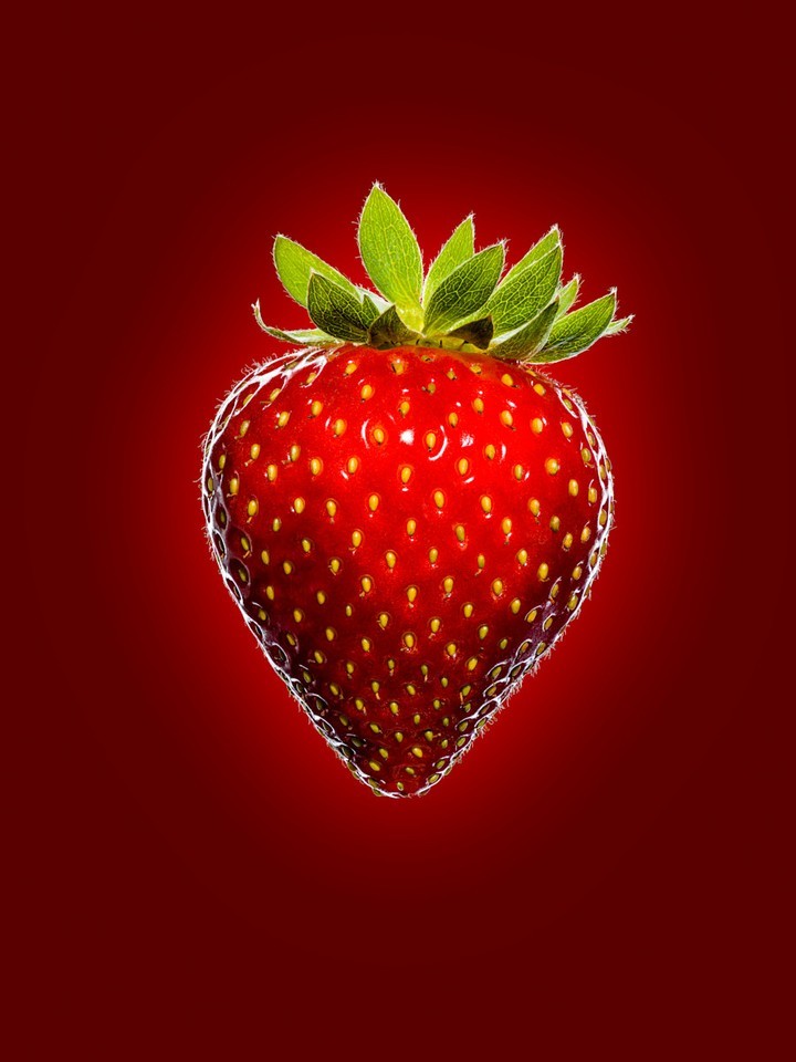 Studio shot of a fresh strawberry with vibrant red color and green leafy top against a dark red backdrop. Close-up on the berry's texture.