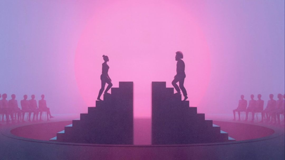Silhouetted figures ascend stairs on either side of a circle stage, with an audience, against a pink and purple gradient background.