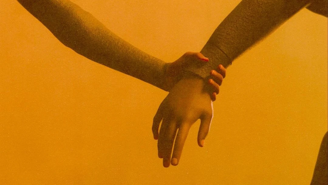 Close-up of two arms and hands in front of a yellow background. One hand is holding the other arm, indicating connection.