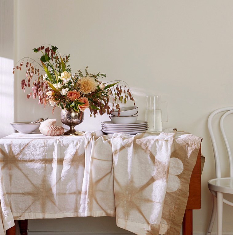 Floral arrangement in silver vase on table with patterned tablecloth alongside plates, shell, pitcher, and chair against neutral wall.