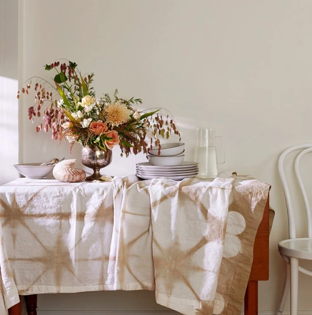 Floral arrangement in silver vase on table with patterned tablecloth alongside plates, shell, pitcher, and chair against neutral wall.