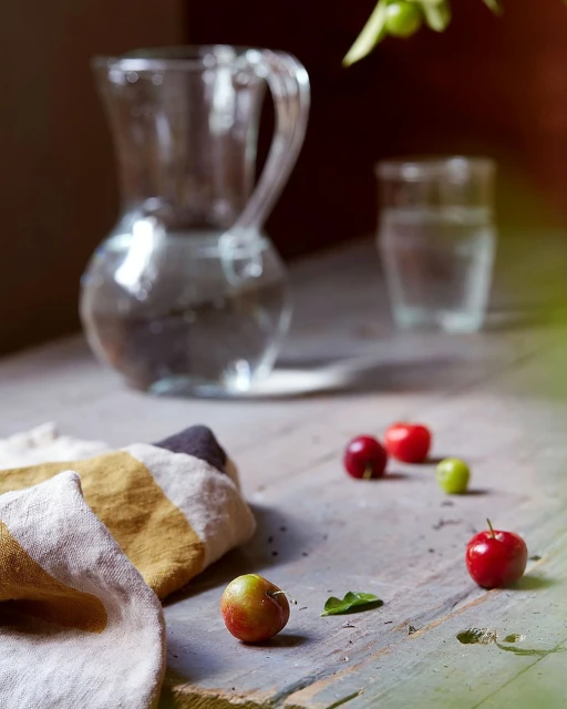 Still life of a glass pitcher and drinking glass with water, surrounded by small red fruits on a weathered wooden table.