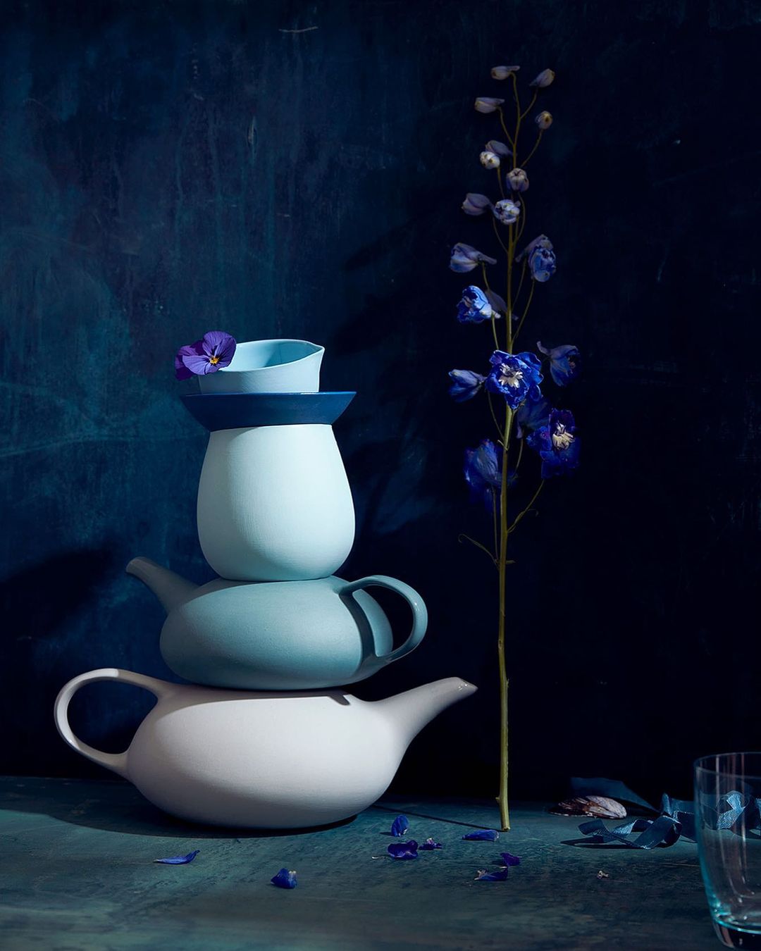 Still life shot of stacked teapots in varying shades of blue and white, accented by a delicate blue flower and scattered petals.
