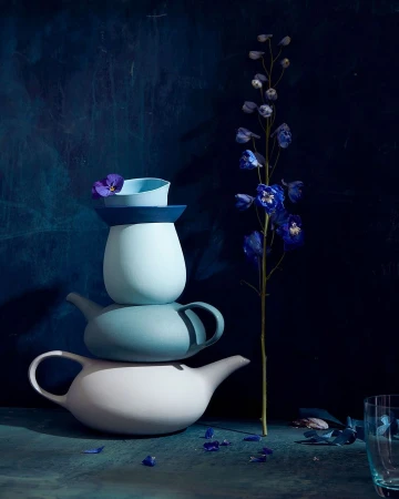Still life shot of stacked teapots in varying shades of blue and white, accented by a delicate blue flower and scattered petals.