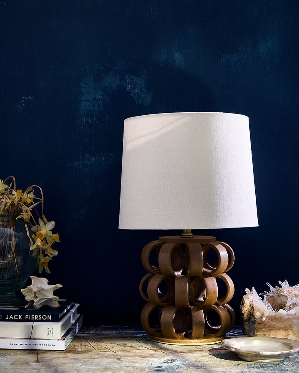 A stylish table lamp with a brown ceramic base and white lampshade sits on a wooden table against a dark blue wall in an interior setting.