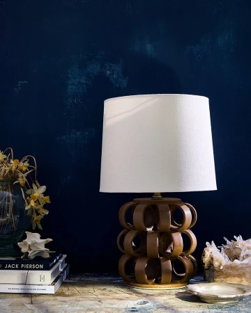A stylish table lamp with a brown ceramic base and white lampshade sits on a wooden table against a dark blue wall in an interior setting.