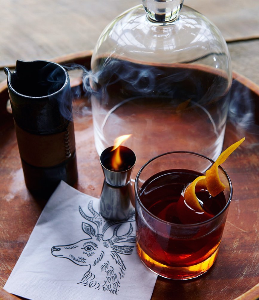 Smoked old fashioned cocktail in a rocks glass with an orange twist, served on a wooden tray with decanter, jigger, and deer napkin.
