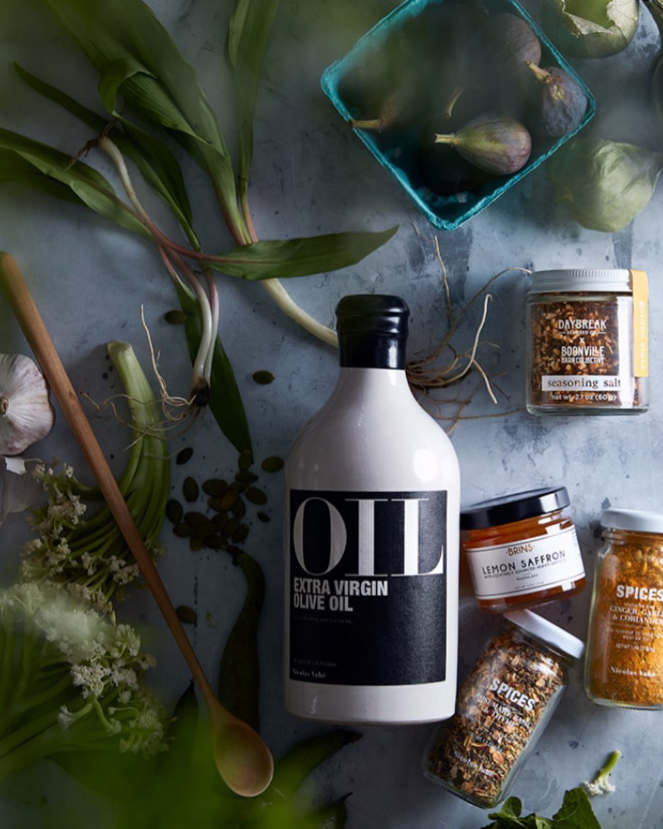 Overhead shot of OLI extra virgin olive oil bottle surrounded by figs, garlic, spices, a wooden spoon, and greenery on a mottled gray surface.