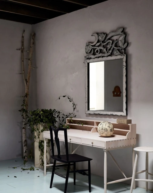 Eclectic interior with light pink desk, black chair, ornate mirror, plants, and white stool in room with light blue floor.